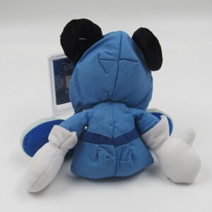 May include: A blue plush toy of a character with black ears and white gloves. The toy is wearing a blue outfit with a matching hood. The toy is sitting on a white surface. A clear plastic case is behind the toy.
