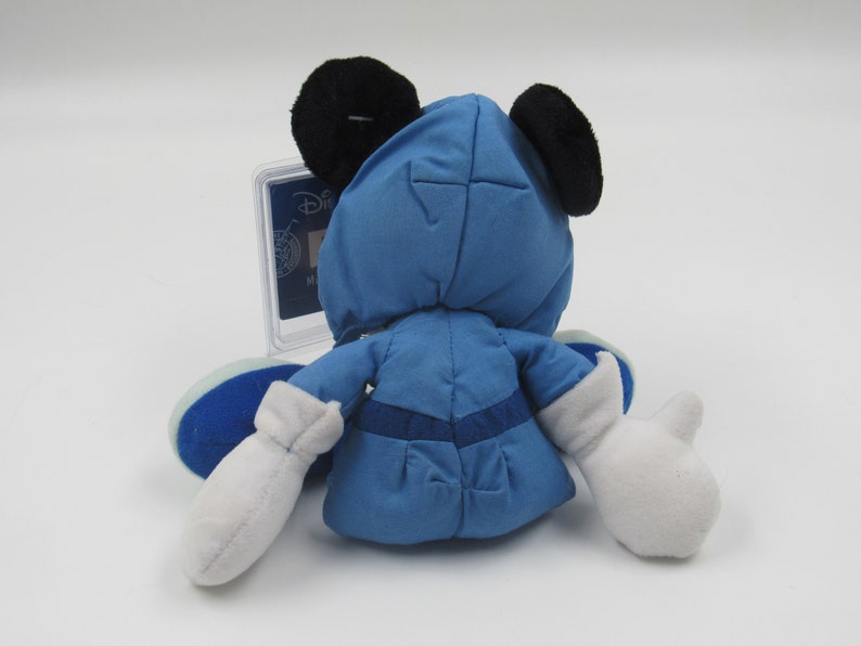 May include: A blue plush toy of a character with black ears and white gloves. The toy is wearing a blue outfit with a matching hood. The toy is sitting on a white surface. A clear plastic case is behind the toy.