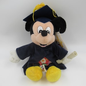 Puede incluir: Un peluche de Mickey Mouse con una túnica de graduación azul y un gorro con un borla amarilla. Está sosteniendo un rollo en su mano derecha.