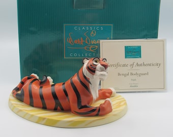 Può includere: Una statuina della collezione Disney Classics di Rajah, la tigre del Bengala di Aladdin. La statuina è arancione con strisce nere, dettagli bianchi e giace su una base gialla. Un certificato di autenticità e una scatola verde acqua sono sullo sfondo.