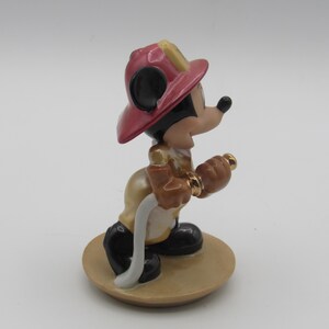Op de afbeelding: Een keramieken beeldje van Mickey Mouse als brandweerman. Mickey draagt een rode helm, een beige jas, een zwarte broek en zwarte schoenen. Hij houdt een goudkleurige spuitmond en een witte slang vast. Het beeldje staat op een beige, ronde basis.