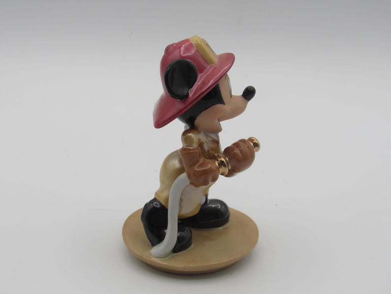Op de afbeelding: Een keramieken beeldje van Mickey Mouse als brandweerman. Mickey draagt een rode helm, een beige jas, een zwarte broek en zwarte schoenen. Hij houdt een goudkleurige spuitmond en een witte slang vast. Het beeldje staat op een beige, ronde basis.
