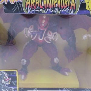 May include: A red and purple action figure of a monster with wings and spider legs. The figure is in a plastic box with a yellow base and the words "Arachniphobia" on the box.