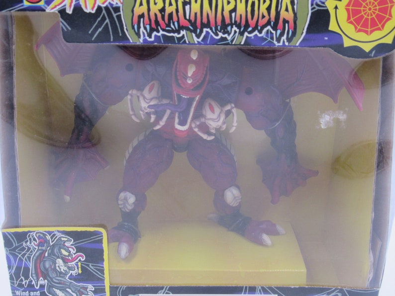 May include: A red and purple action figure of a monster with wings and spider legs. The figure is in a plastic box with a yellow base and the words "Arachniphobia" on the box.