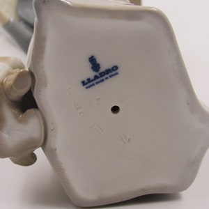 May include: A ceramic figurine with a light grey base and a small brown animal figure. The base has the text "LLADRO HAND MADE IN SPAIN" in blue. The figurine is likely a collectible item.