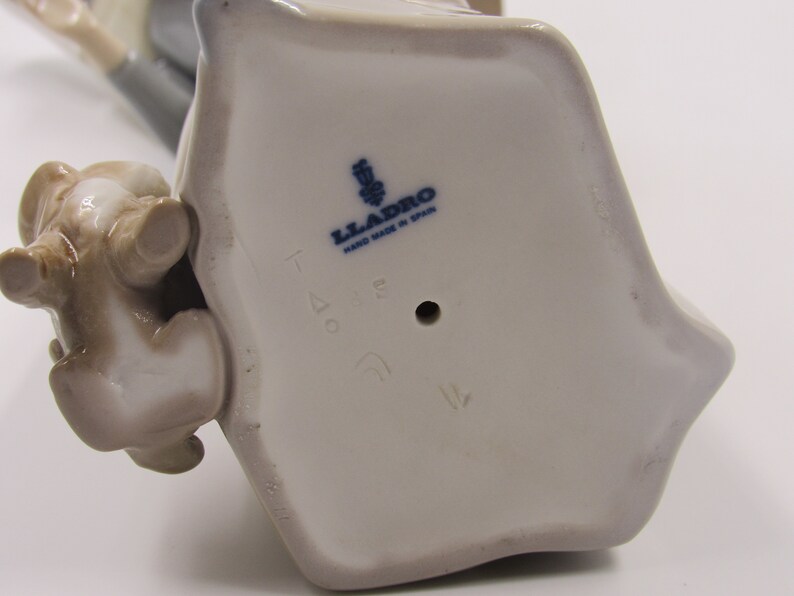 May include: A ceramic figurine with a light grey base and a small brown animal figure. The base has the text "LLADRO HAND MADE IN SPAIN" in blue. The figurine is likely a collectible item.