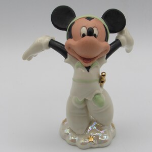 Op de afbeelding: Een keramieken beeldje van Mickey Mouse in een witte outfit met groene accenten, zwarte oren en een grote roze neus. Het beeldje staat op een wolk-vormige basis met gouden sterren.