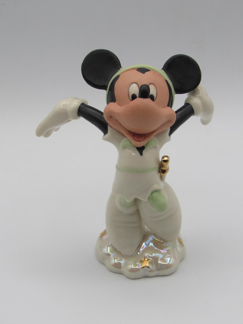 Op de afbeelding: Een keramieken beeldje van Mickey Mouse in een witte outfit met groene accenten, zwarte oren en een grote roze neus. Het beeldje staat op een wolk-vormige basis met gouden sterren.