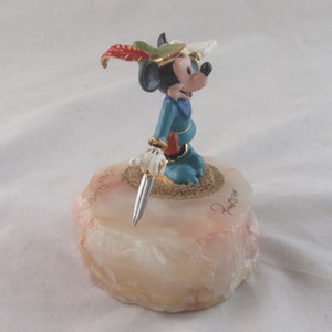 K&ouml;nnte beinhalten: Eine Mickey Mouse-Figur in blauem Outfit mit Federhut und silbernem Dolch. Die Figur steht auf einem hellrosa und wei&szlig;en Steinsockel mit goldenen Akzenten und einer Signatur. Der Sockel ist ca. 5 cm hoch.