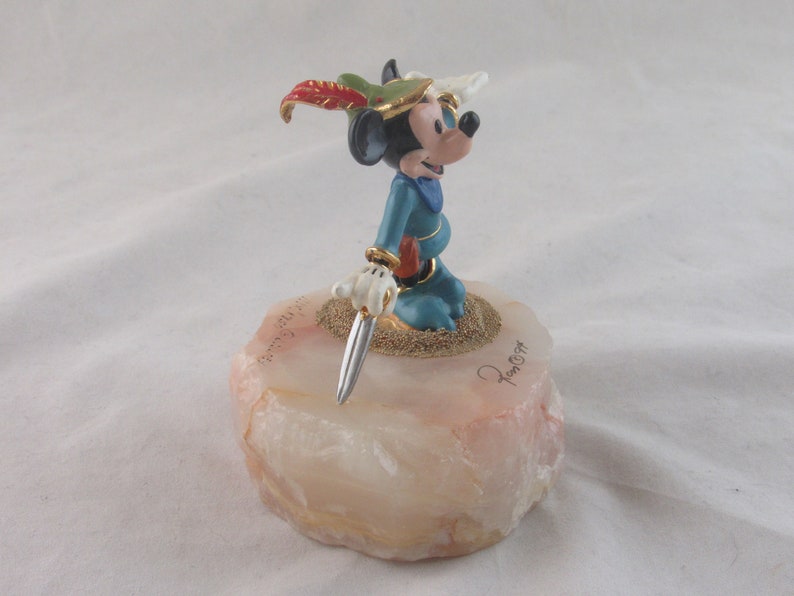 K&ouml;nnte beinhalten: Eine Mickey Mouse-Figur in blauem Outfit mit Federhut und silbernem Dolch. Die Figur steht auf einem hellrosa und wei&szlig;en Steinsockel mit goldenen Akzenten und einer Signatur. Der Sockel ist ca. 5 cm hoch.