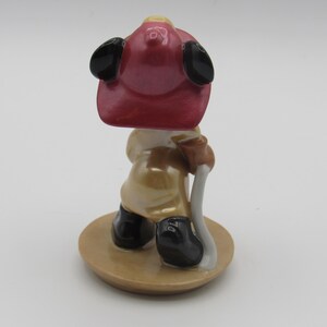 Op de afbeelding: Een keramieken beeldje van een brandweerman, van achteren gezien. Het figuur draagt een rode helm met zwarte oorflappen, een beige uniform en zwarte laarzen. Het houdt een wit en bruin gereedschap vast en staat op een ronde, beige basis.