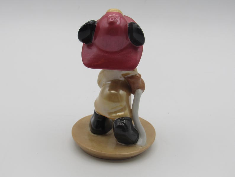 Op de afbeelding: Een keramieken beeldje van een brandweerman, van achteren gezien. Het figuur draagt een rode helm met zwarte oorflappen, een beige uniform en zwarte laarzen. Het houdt een wit en bruin gereedschap vast en staat op een ronde, beige basis.