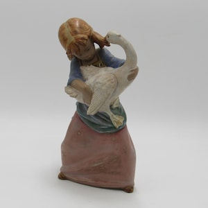 May include: A ceramic figurine depicting a young person holding a white goose. The figure is dressed in a blue top, a green sash, and a pink skirt. The goose has a long neck and is held close to the person's face. The figurine is approximately 6 inches tall.