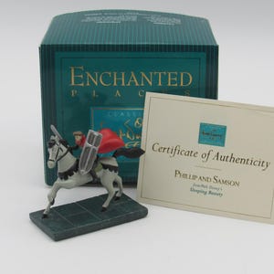 May include: A figurine of a rider on a grey horse with a red cape and silver shield, standing on a dark green base. Behind it is a teal box labelled Enchanted Places and a Certificate of Authenticity for Phillip and Samson from Sleeping Beauty.