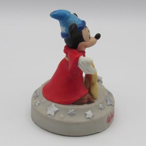 May include: A figurine of Mickey Mouse dressed as a sorcerer, wearing a blue hat and red robe. The figure is standing on a gray base decorated with stars and the word "Walt" in cursive.