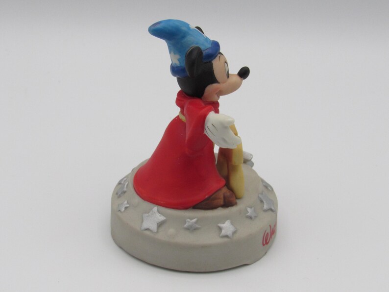 May include: A figurine of Mickey Mouse dressed as a sorcerer, wearing a blue hat and red robe. The figure is standing on a gray base decorated with stars and the word "Walt" in cursive.