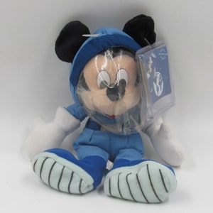 May include: A plush Mickey Mouse toy dressed in a blue astronaut suit with a clear helmet. The toy has black ears, white gloves, and blue and white striped shoes. A clear tag is attached to the toy.
