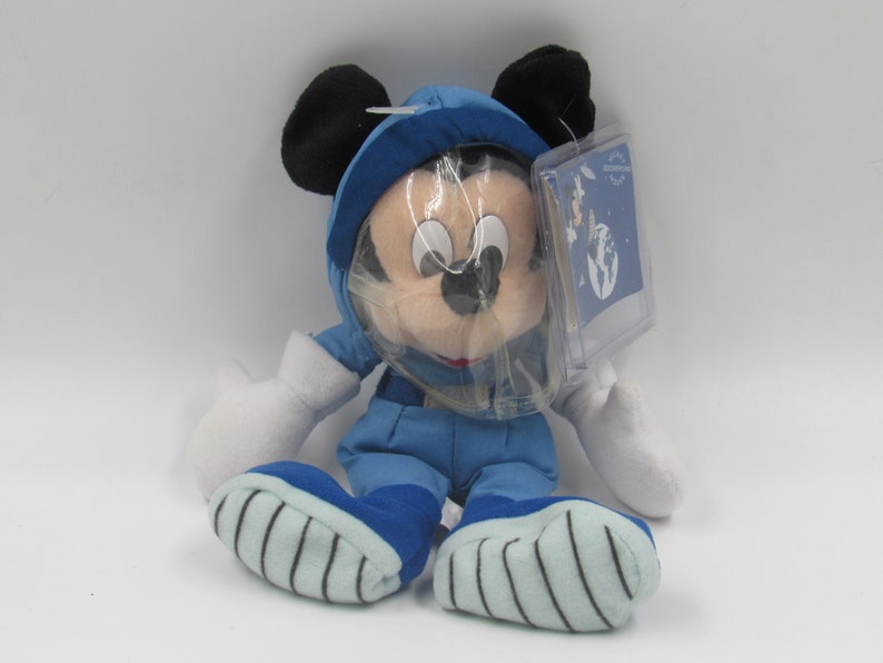 May include: A plush Mickey Mouse toy dressed in a blue astronaut suit with a clear helmet. The toy has black ears, white gloves, and blue and white striped shoes. A clear tag is attached to the toy.