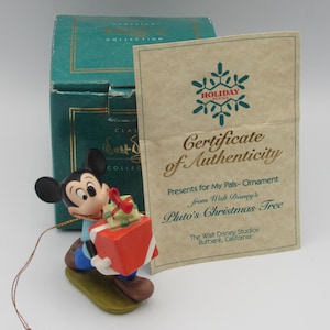 May include: A Mickey Mouse Christmas ornament holding a red gift box with a green bow. The ornament is part of the "Holiday Series" and comes with a certificate of authenticity from Walt Disney Studios, Burbank, California.