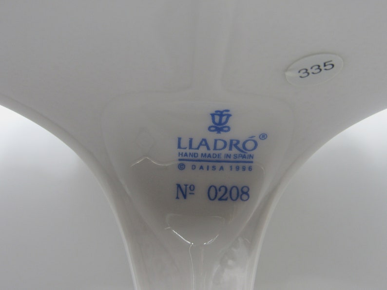May include: A white porcelain figurine with blue markings. The figurine is marked with the Lladro logo, "Hand Made in Spain", "DAISA 1986", and "N&deg; 0208".