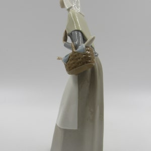 May include: A porcelain figurine of a woman in a long dress, carrying a basket. The figure wears a head covering and a shawl. The dress is a muted grey, and the basket is brown. The figurine is set against a white background.
