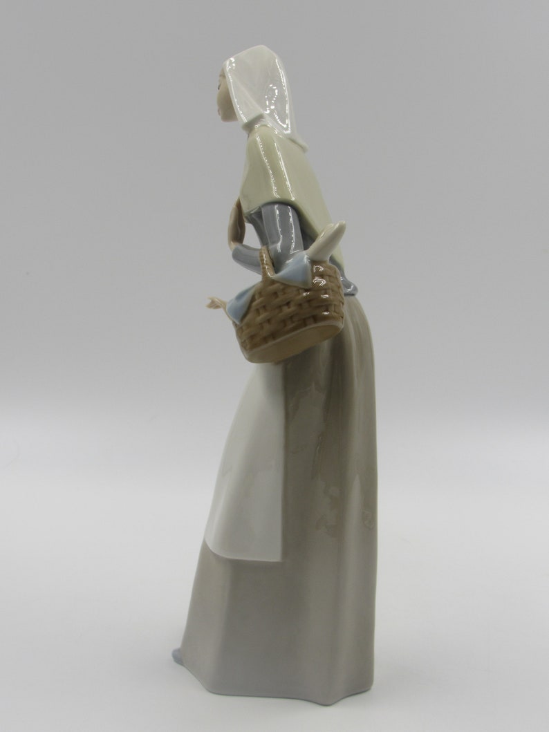 May include: A porcelain figurine of a woman in a long dress, carrying a basket. The figure wears a head covering and a shawl. The dress is a muted grey, and the basket is brown. The figurine is set against a white background.