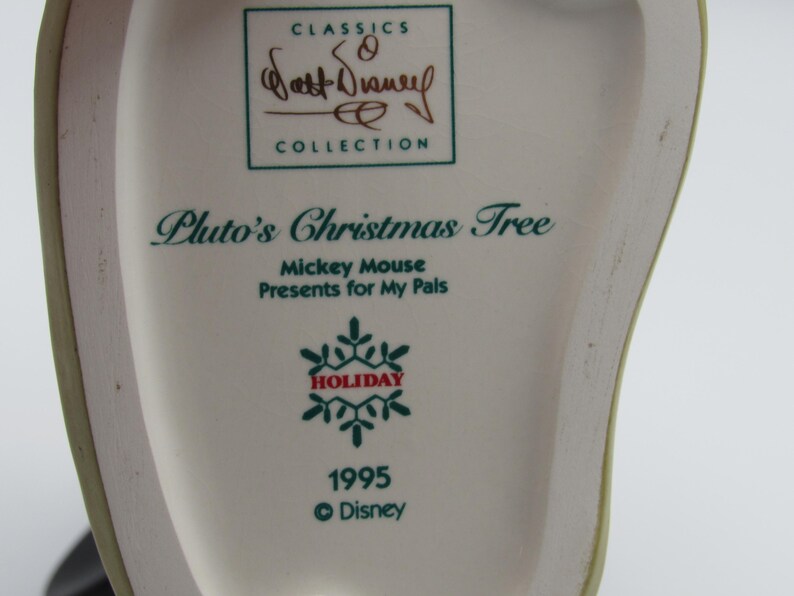 May include: A close-up of a white ceramic collectible featuring text that reads "Pluto's Christmas Tree" and "Mickey Mouse Presents for My Pals." The design includes a snowflake and the year 1995, with the Disney logo. The item is part of the "Walt Disney Collection."