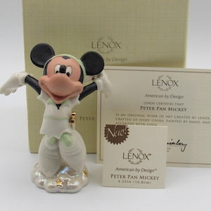 Op de afbeelding: Een Lenox Peter Pan Mickey beeldje, 10,8 cm hoog, in een witte outfit met groene accenten, staand op een wolkenbasis. Het beeldje heeft zwarte oren en een roze gezicht. De doos en het certificaat zijn zichtbaar.