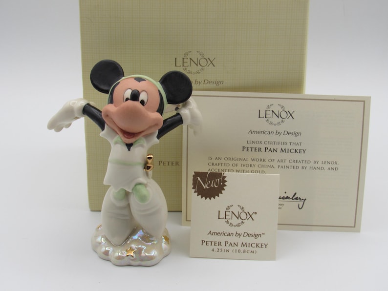 Op de afbeelding: Een Lenox Peter Pan Mickey beeldje, 10,8 cm hoog, in een witte outfit met groene accenten, staand op een wolkenbasis. Het beeldje heeft zwarte oren en een roze gezicht. De doos en het certificaat zijn zichtbaar.