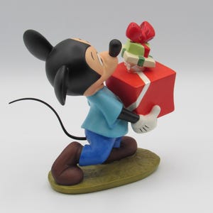 May include: A figurine of Mickey Mouse, wearing a blue shirt, blue pants, and brown shoes, carrying a large red gift box with a white ribbon and smaller green and red gifts. The figurine is on a green base.
