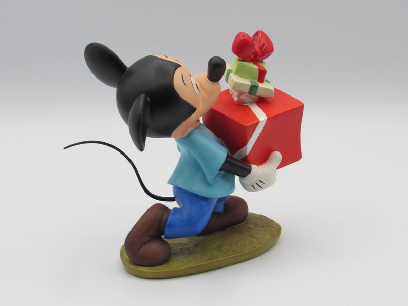 May include: A figurine of Mickey Mouse, wearing a blue shirt, blue pants, and brown shoes, carrying a large red gift box with a white ribbon and smaller green and red gifts. The figurine is on a green base.