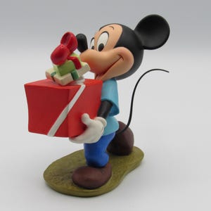 May include: A Mickey Mouse figurine holding a large red gift box with a white ribbon and smaller gifts on top. Mickey is wearing a blue shirt, blue pants, white gloves, and brown shoes. The figurine is on a green and brown base.