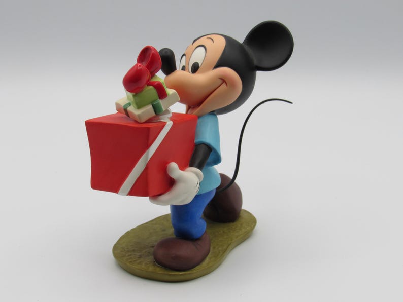 May include: A Mickey Mouse figurine holding a large red gift box with a white ribbon and smaller gifts on top. Mickey is wearing a blue shirt, blue pants, white gloves, and brown shoes. The figurine is on a green and brown base.