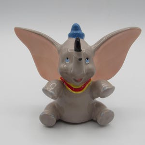 May include: A ceramic figurine of Dumbo the elephant, featuring large pink ears, a blue hat, and a red and yellow collar. The figure is in a seated position with its arms outstretched, and has a smiling expression.