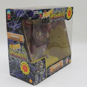 May include: A red and purple plastic toy figure of a vampire spider from the Spider-Man Arachnophobia toy line. The figure has a wind-up action and is packaged in a clear plastic box with a black and green background. The box has the words "Spider-Man Arachnophobia" and "Vampider" printed on it.