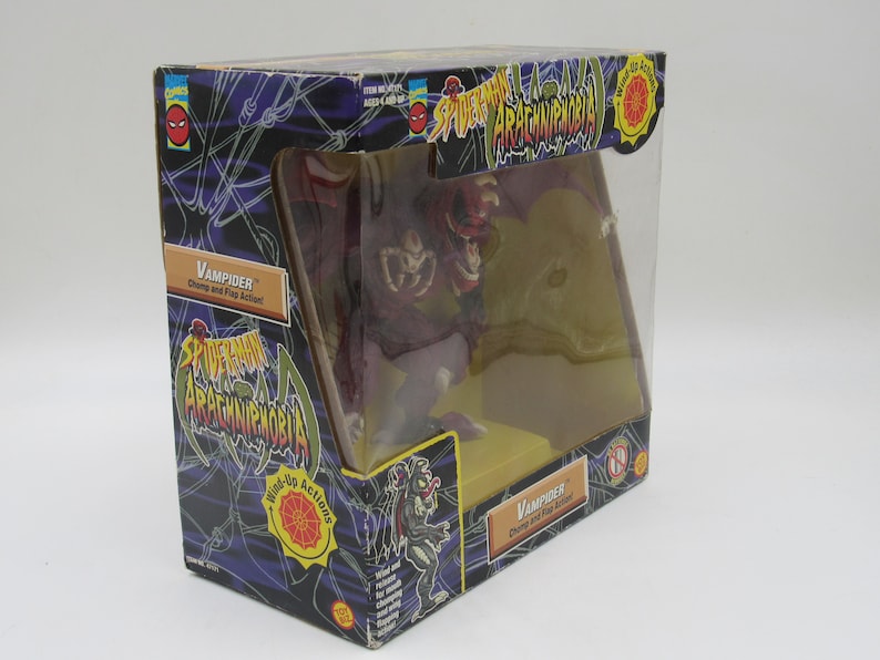 May include: A red and purple plastic toy figure of a vampire spider from the Spider-Man Arachnophobia toy line. The figure has a wind-up action and is packaged in a clear plastic box with a black and green background. The box has the words "Spider-Man Arachnophobia" and "Vampider" printed on it.