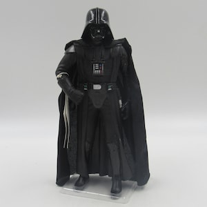 May include: A black Darth Vader action figure with a black cape and a white belt. The figure is standing on a clear plastic base.