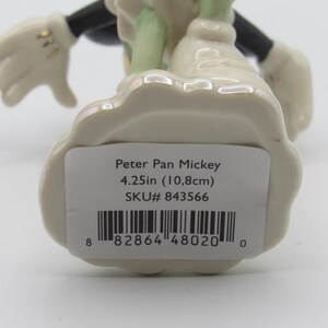 Op de afbeelding: Een keramieken beeldje van Peter Pan Mickey Mouse, met een cr&egrave;mekleurige basis en accenten. Het beeldje is 10,8 cm hoog. De figuur heeft zwarte armen en benen, witte handschoenen en groene kleding.
