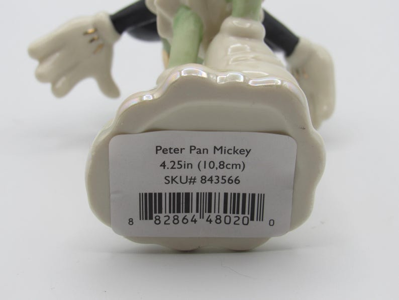 Op de afbeelding: Een keramieken beeldje van Peter Pan Mickey Mouse, met een cr&egrave;mekleurige basis en accenten. Het beeldje is 10,8 cm hoog. De figuur heeft zwarte armen en benen, witte handschoenen en groene kleding.