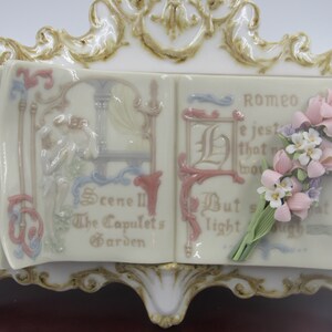 May include: A porcelain figurine of a book with a floral bouquet. The book is open and the pages are decorated with a scene from Romeo and Juliet. The text on the pages reads "Scene III The Capulet's Garden" and "Romeo He jesteth that which wor But soft what light through yonder window breaks"