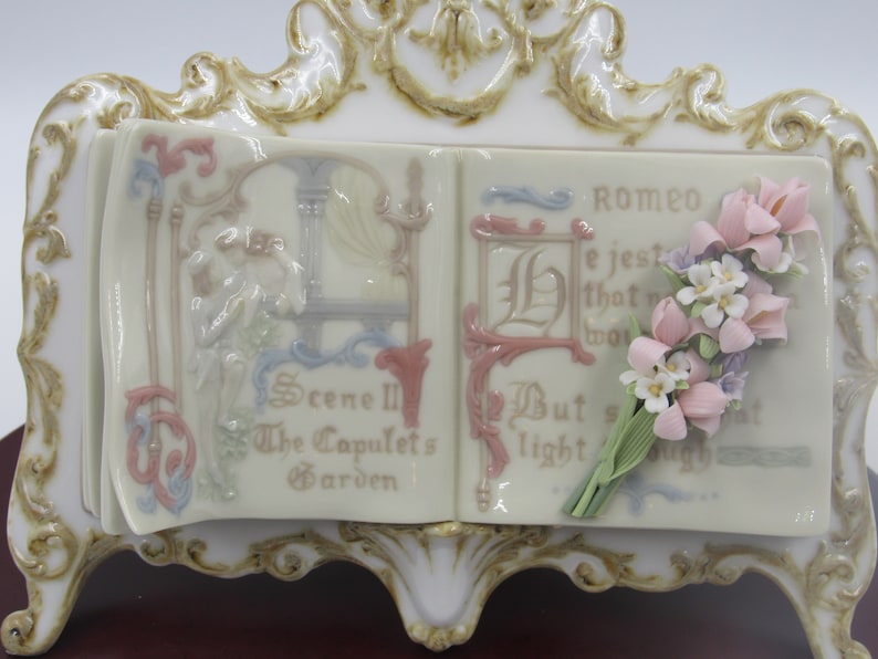 May include: A porcelain figurine of a book with a floral bouquet. The book is open and the pages are decorated with a scene from Romeo and Juliet. The text on the pages reads "Scene III The Capulet's Garden" and "Romeo He jesteth that which wor But soft what light through yonder window breaks"