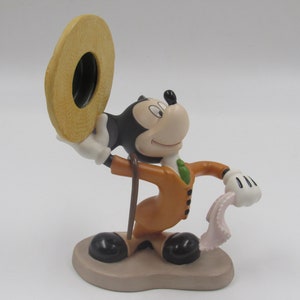 May include: A figurine of Mickey Mouse in a tan suit, holding a hat and a handkerchief. The figure has black ears, a white face, and black shoes. The base is a light brown color.