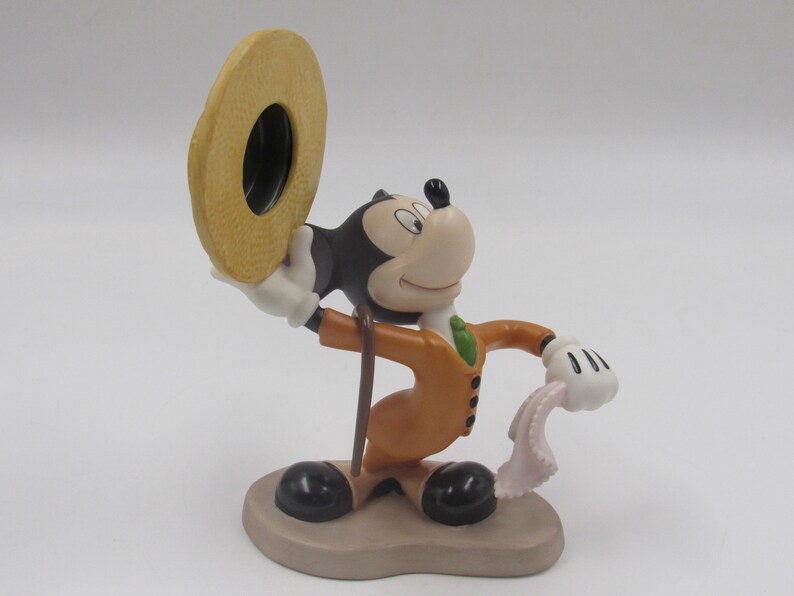May include: A figurine of Mickey Mouse in a tan suit, holding a hat and a handkerchief. The figure has black ears, a white face, and black shoes. The base is a light brown color.