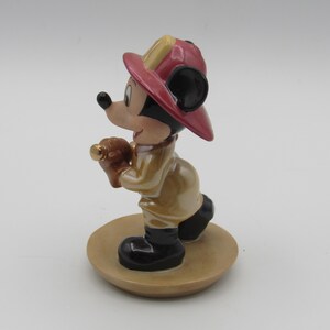 Op de afbeelding: Een keramieken beeldje van Mickey Mouse als brandweerman. Mickey draagt een rode en gele brandweerhelm, een beige jas en zwarte laarzen. Hij houdt een bruin voorwerp met een gouden punt vast. Het beeldje staat op een beige, ronde basis.