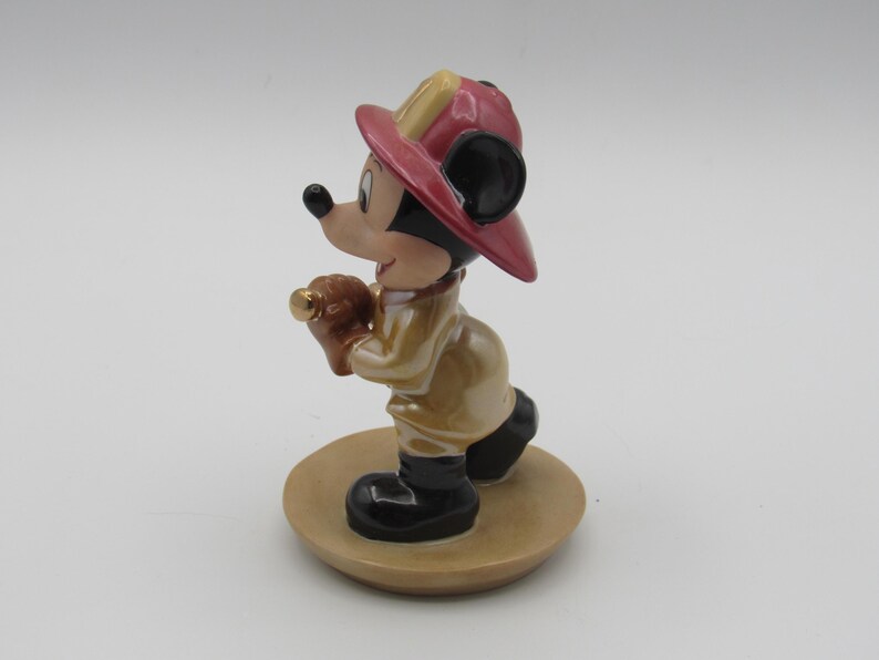 Op de afbeelding: Een keramieken beeldje van Mickey Mouse als brandweerman. Mickey draagt een rode en gele brandweerhelm, een beige jas en zwarte laarzen. Hij houdt een bruin voorwerp met een gouden punt vast. Het beeldje staat op een beige, ronde basis.