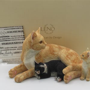 May include: A Lenox figurine set featuring a mother cat and two kittens. The mother cat is orange and white, with a black and white kitten nestled beneath her. A smaller, tan kitten sits nearby. The set is displayed in front of a box.