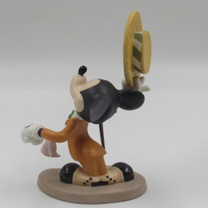 May include: A figurine of Mickey Mouse in a brown suit, holding a hat and a handkerchief. The figure has black ears, a black head, and white gloves. The hat is yellow with green stripes.