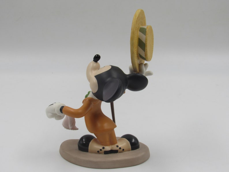 May include: A figurine of Mickey Mouse in a brown suit, holding a hat and a handkerchief. The figure has black ears, a black head, and white gloves. The hat is yellow with green stripes.