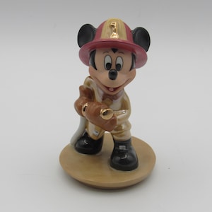 Op de afbeelding: Een keramieken beeldje van Mickey Mouse verkleed als brandweerman. Mickey draagt een rode en gouden helm met het nummer 1, een beige uniform en zwarte laarzen. Hij houdt een bruine brandslang vast en staat op een beige cirkelvormige basis.