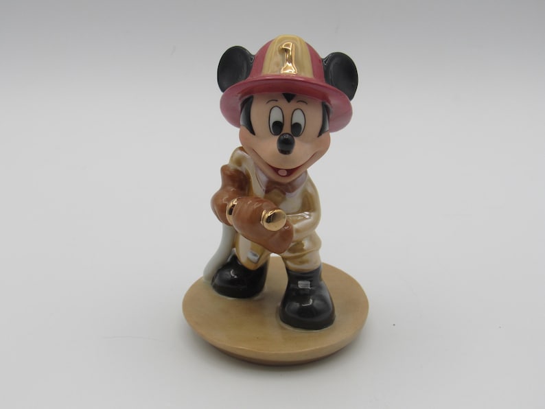Op de afbeelding: Een keramieken beeldje van Mickey Mouse verkleed als brandweerman. Mickey draagt een rode en gouden helm met het nummer 1, een beige uniform en zwarte laarzen. Hij houdt een bruine brandslang vast en staat op een beige cirkelvormige basis.